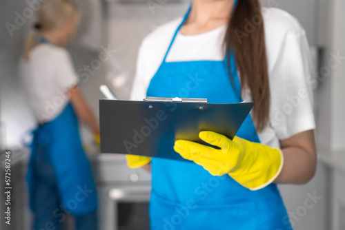 Close-up of a young cleaning woman in an apron writes down a report on the work done on a paper tablet against the background of a blurred employee. Cleaning company in the kitchen  teamwork