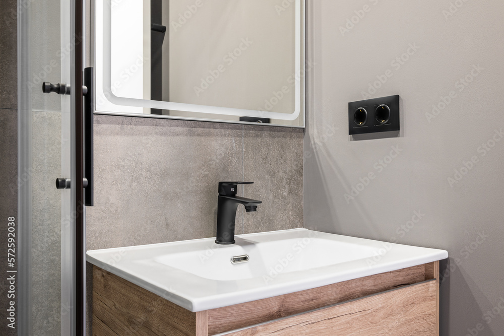 Closeup of a white square ceramic sink on a wooden vanity. Fittings and faucet made of ferrous metal. On the wall is a designer mirror with a white frame. Walls in gray pleasant tones. Closeup of a white square ceramic sink on a wooden vanity. Fittings and faucet made of ferrous metal. On the wall is a designer mirror with a white frame. Walls in gray pleasant tones.