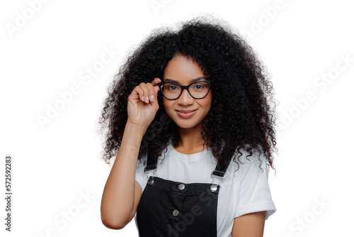 Pretty African American woman keeps hand on frame of glasses, smiles happily, wears white casual t shirt and black overalls, enjoys spare time, poses over violet background. Ethnicity, fashion concept