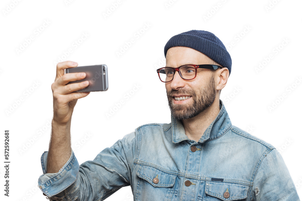 Pleased smiling bearded male with happy expression poses at camera of smart phone, takes photo of himself, wears denim jacket and glasses, isolated over white background. People and technology concept Pleased smiling bearded male with happy expression poses at camera of smart phone, takes photo of himself, wears denim jacket and glasses, isolated over white background. People and technology concept