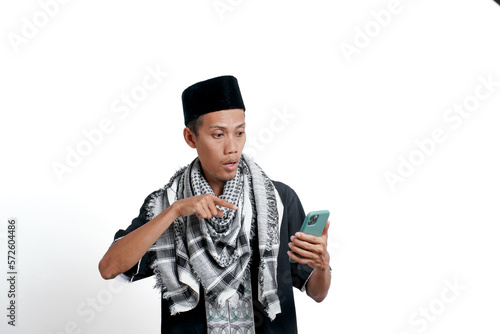 A religious Muslim Asian man wearing a turban, Muslim clothes and a cap, holding a smartphone like he's on a video call, and is shocked. Isolated on white background. photo
