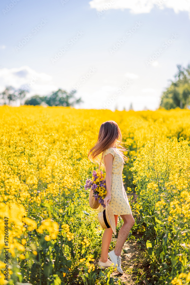 Fototapeta premium girl in a hat with a bag of flowers walks in a field of rapeseed. Yellow flower field. View from the back