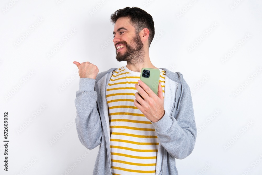 caucasian man wearing casual sportswear over white wall using and texting with smartphone pointing and showing with thumb up to the side with happy face smiling caucasian man wearing casual sportswear over white wall using and texting with smartphone pointing and showing with thumb up to the side with happy face smiling