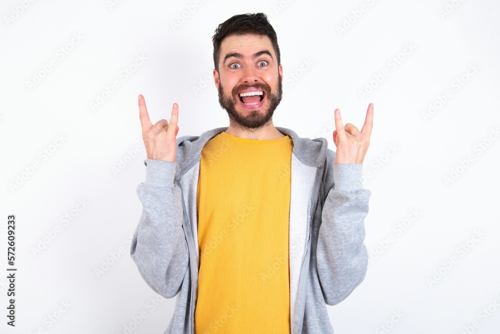Fototapeta premium caucasian man wearing casual sportswear over white wall makes rock n roll sign looks self confident and cheerful enjoys cool music at party. Body language concept.