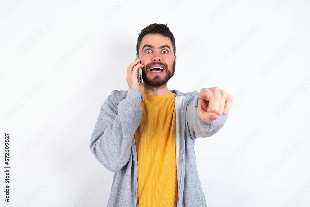 Fototapeta premium Positive caucasian man wearing casual sportswear over white wall indicates directly at camera has telephone conversation smiles broadly enjoys talking long hours. You join me