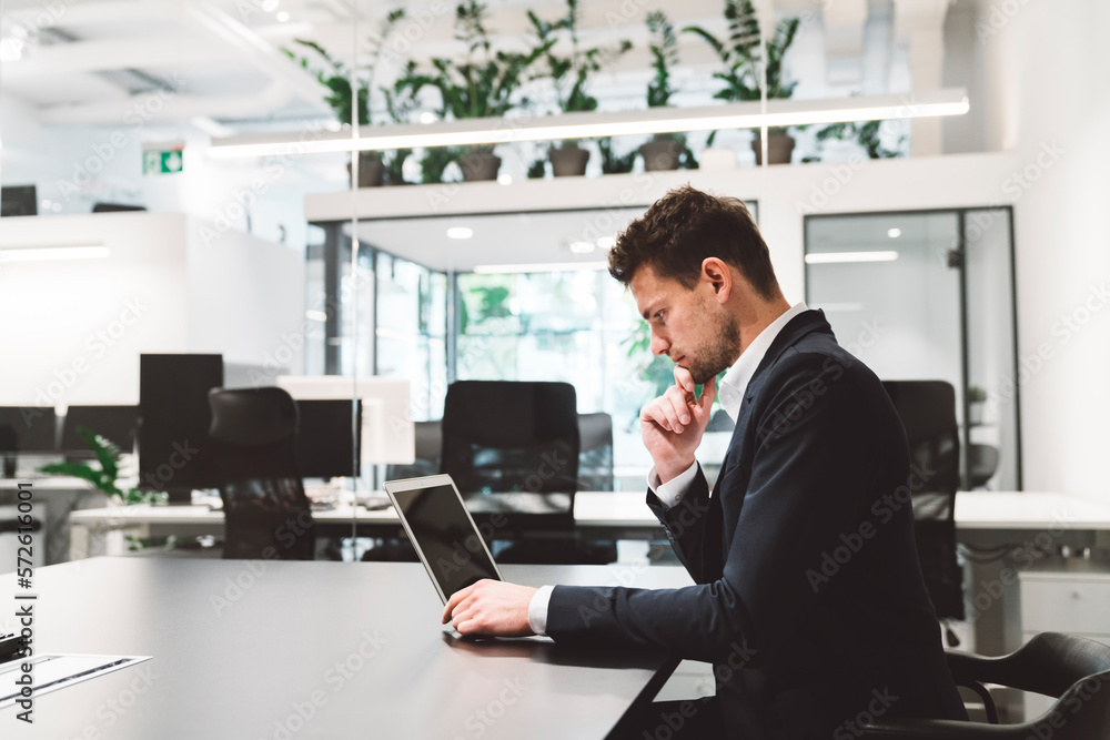 Side view of caucasian businessman in black suit working on his laptop in modern bright offices Side view of caucasian businessman in black suit working on his laptop in modern bright offices