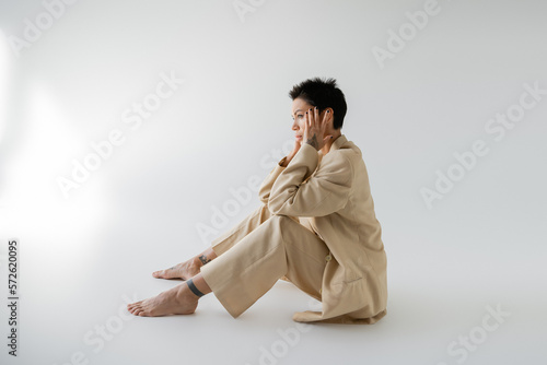 side view of tattooed barefoot woman in beige pantsuit sitting with hands near face on grey background.