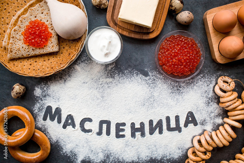 Russian traditions. Russian holiday Maslenitsa. A stack of pancakes, red caviar, bagels, bagels, a wooden spoon, eggs and butter. The Russian phrase Maslenitsa is written with flour. View from above.