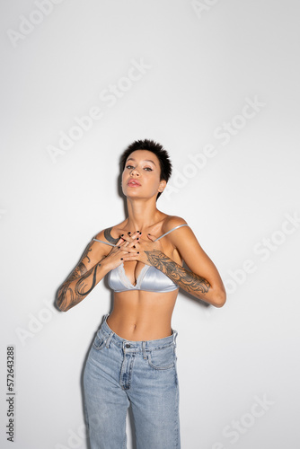 sexy tattooed woman in silk bra and jeans holding hands on chest while looking at camera on grey background.