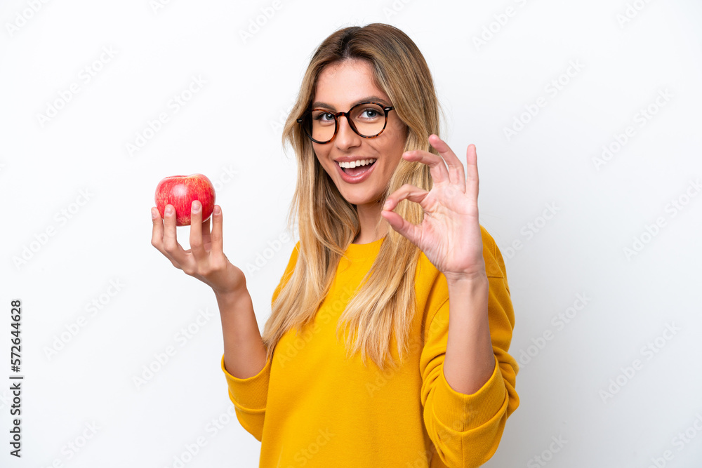 Young Uruguayan woman with an apple isolated on white background showing ok sign with fingers Young Uruguayan woman with an apple isolated on white background showing ok sign with fingers