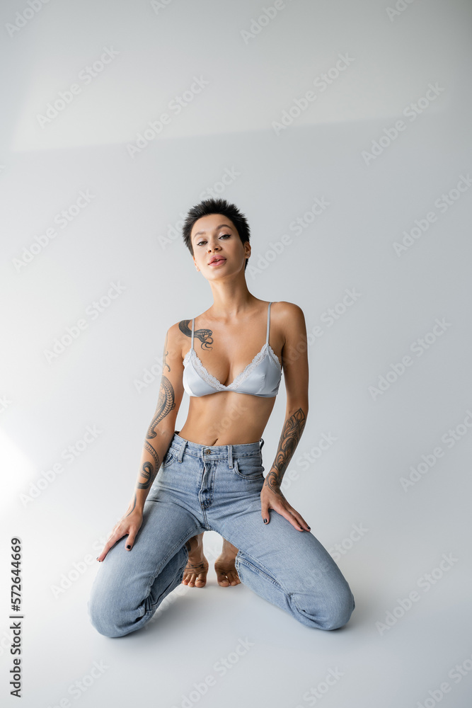 sensual brunette woman in satin bralette and jeans posing on knees on grey background.