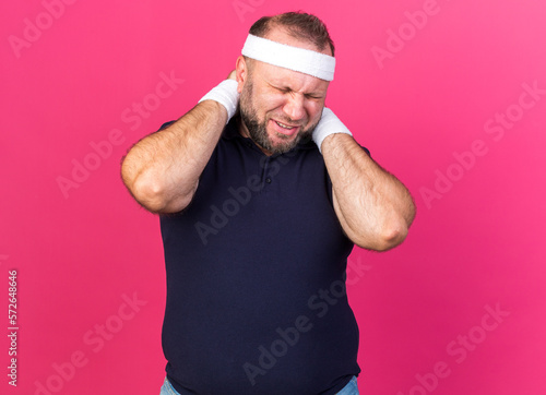 aching adult slavic sporty man wearing headband and wristbands holding his neck with hands isolated on pink background with copy space © HN Works