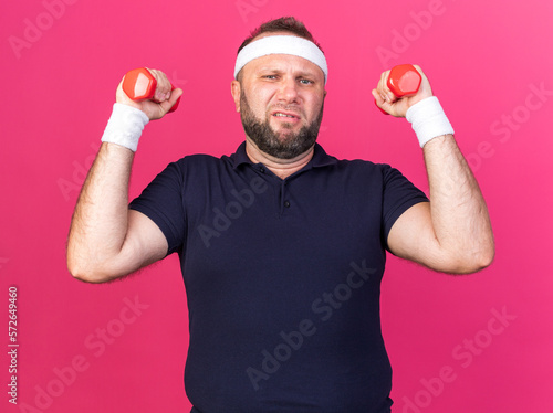 unpleased adult slavic sporty man wearing headband and wristbands holding dumbbells isolated on pink background with copy space photo