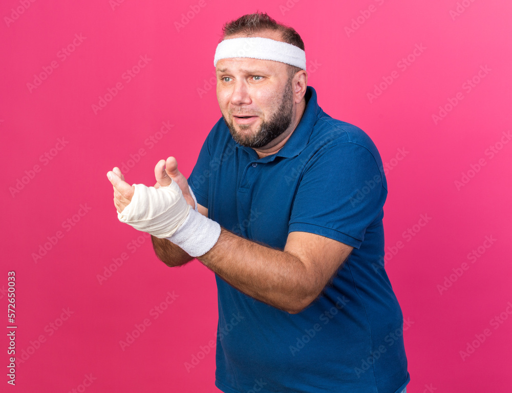 sad adult slavic sporty man wearing headband and wristbands looking and pointing at side isolated on pink background with copy space sad adult slavic sporty man wearing headband and wristbands looking and pointing at side isolated on pink background with copy space