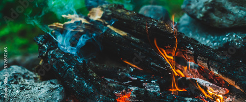 Vivid smoldered firewoods burned in fire close-up. Atmospheric warm background with orange flame of campfire and blue smoke. Wonderful full frame image of bonfire. Burning logs in beautiful fire.