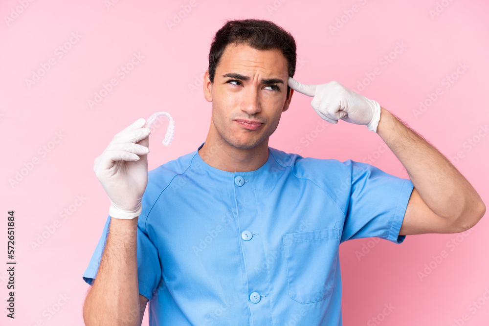 Dentist caucasian man holding invisible braces isolated on pink background having doubts and thinking Dentist caucasian man holding invisible braces isolated on pink background having doubts and thinking