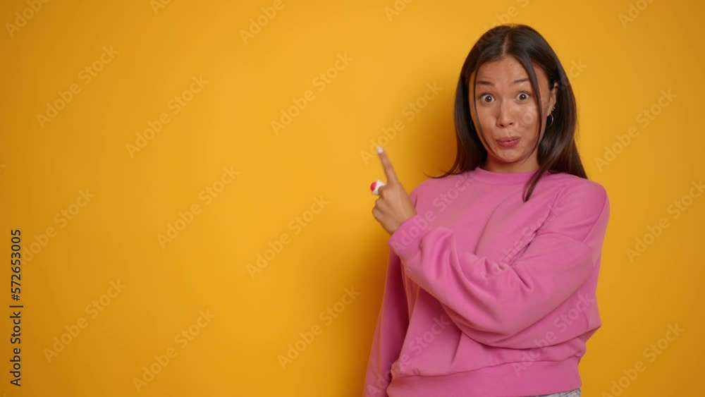 Happy fun young woman 20s wearing casual pink hoodie pointing index fingers aside on mock up copy space area spreading hands isolated over yellow background Happy fun young woman 20s wearing casual pink hoodie pointing index fingers aside on mock up copy space area spreading hands isolated over yellow background