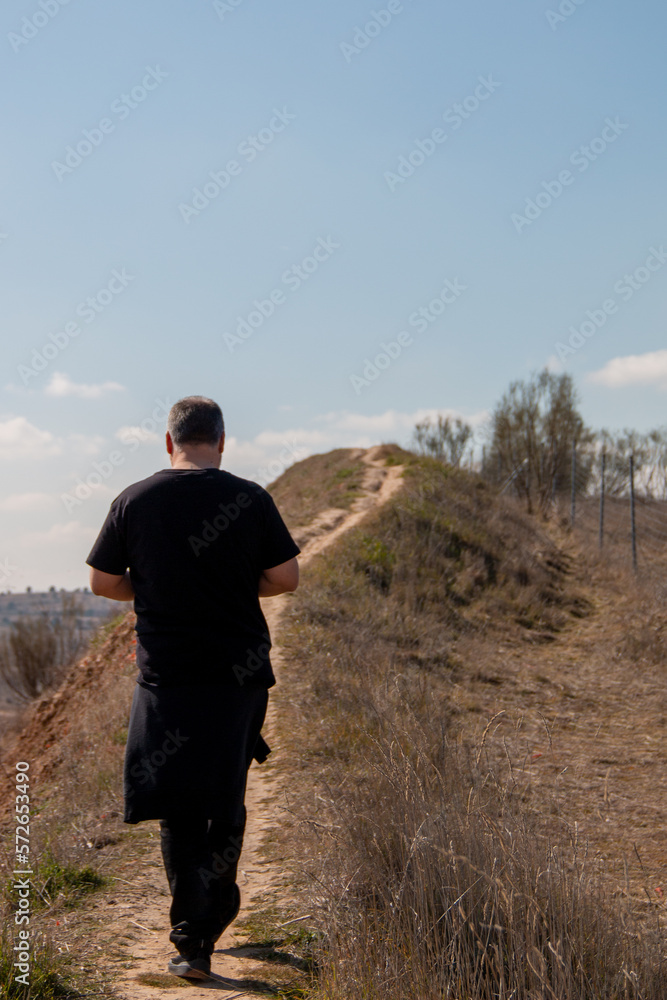 Man dressed in black walking on a path on a cliff where you can see the blue sky