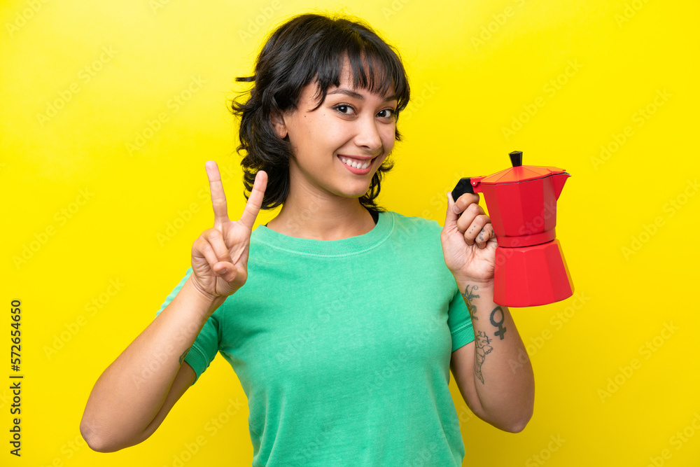 Young Argentinian woman holding coffee pot isolated on yellow background smiling and showing victory sign Young Argentinian woman holding coffee pot isolated on yellow background smiling and showing victory sign