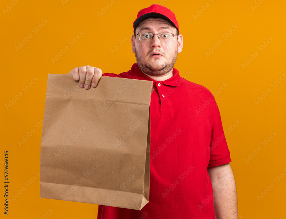anxious overweight young deliveryman in optical glasses holding paper package isolated on orange background with copy space