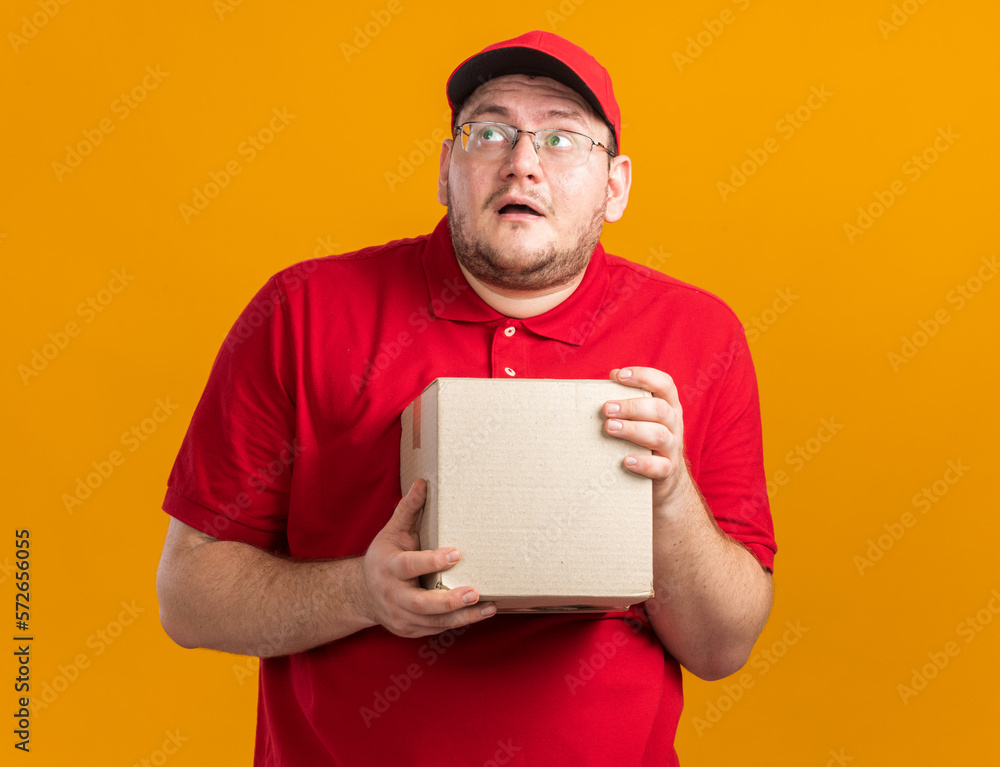 surprised overweight young deliveryman in optical glasses holding cardboard box and looking at side isolated on orange background with copy space surprised overweight young deliveryman in optical glasses holding cardboard box and looking at side isolated on orange background with copy space