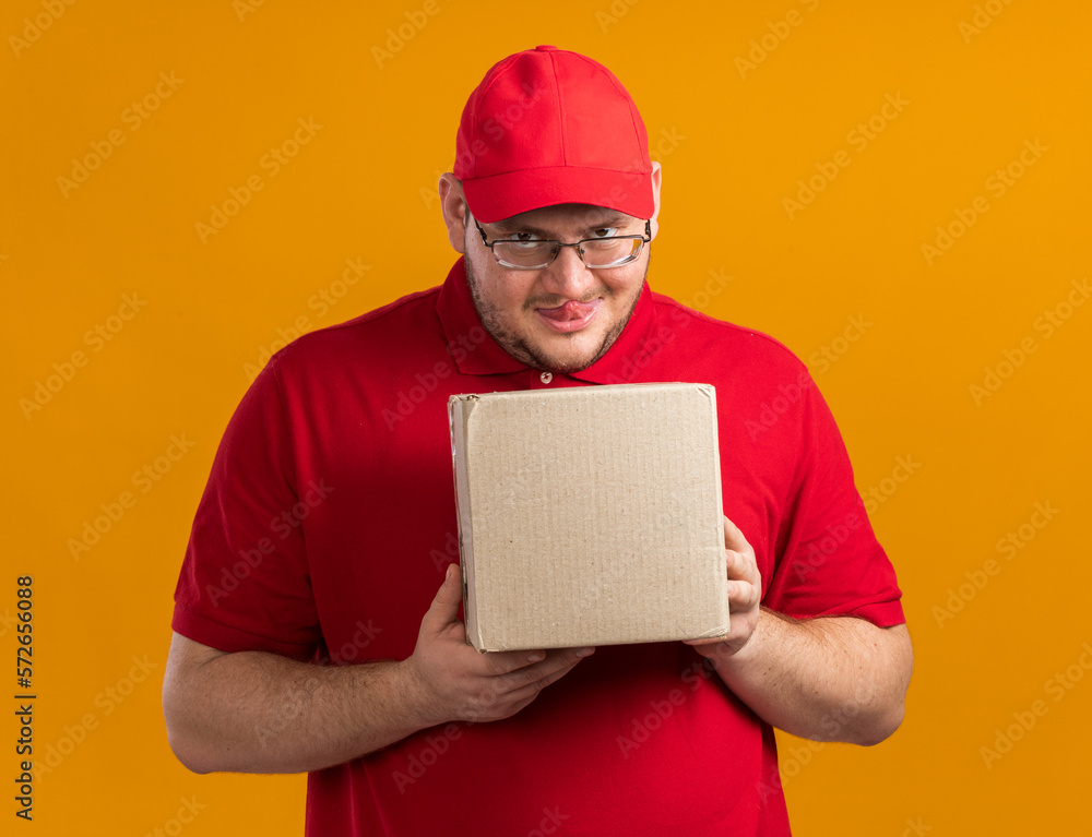 evil joyful overweight young deliveryman in optical glasses stucks out tongue holding cardboard box isolated on orange background with copy space evil joyful overweight young deliveryman in optical glasses stucks out tongue holding cardboard box isolated on orange background with copy space