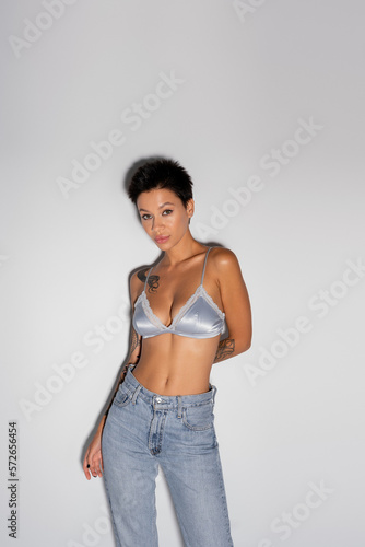 tattooed brunette woman in silk bralette and blue jeans standing with hand behind back on grey background.