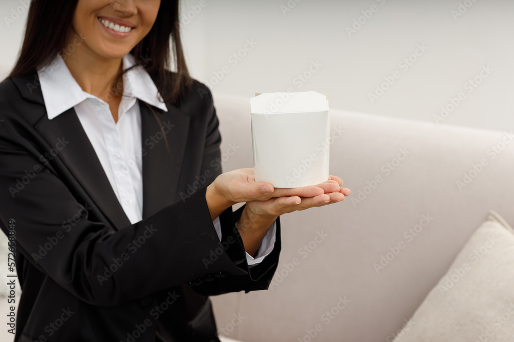 Happy pretty young woman in formal suit and white shirt, sitting on the sofa, stretches out her hands to receive a paper box with food. Fast food during the lunch break in the office. Food delivery. Happy pretty young woman in formal suit and white shirt, sitting on the sofa, stretches out her hands to receive a paper box with food. Fast food during the lunch break in the office. Food delivery.