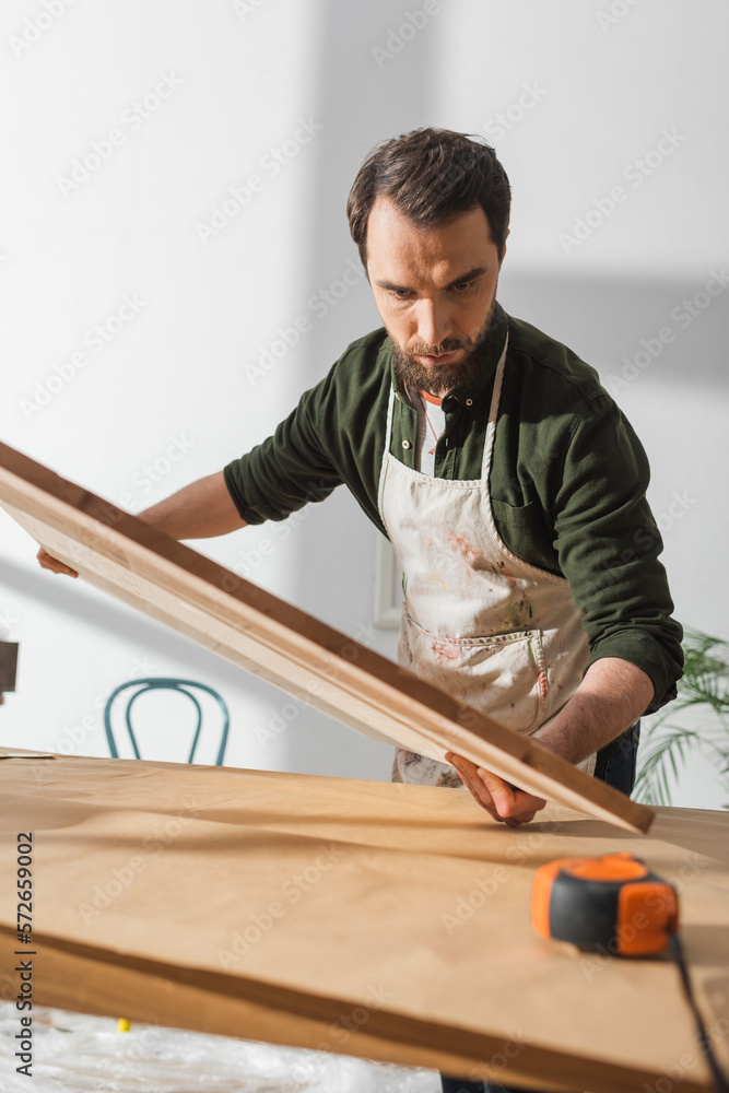 Bearded craftsman in apron holding wooden board near ruler in workshop.