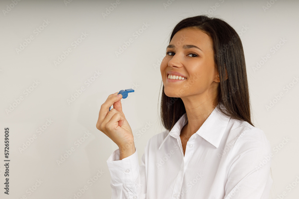 Portrait of attractive smiling girl brunette holding blue whistle in her hand and looking at the camera. Happy young woman with long hair in white shirt on isolated background. Fan, coach, counselor. Portrait of attractive smiling girl brunette holding blue whistle in her hand and looking at the camera. Happy young woman with long hair in white shirt on isolated background. Fan, coach, counselor.