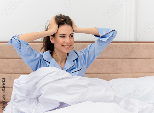 young beautiful woman in blue pajamas sitting in bed happy and positive smiling cheerfully touching her head waking up in bedroom interior on light background © HN Works