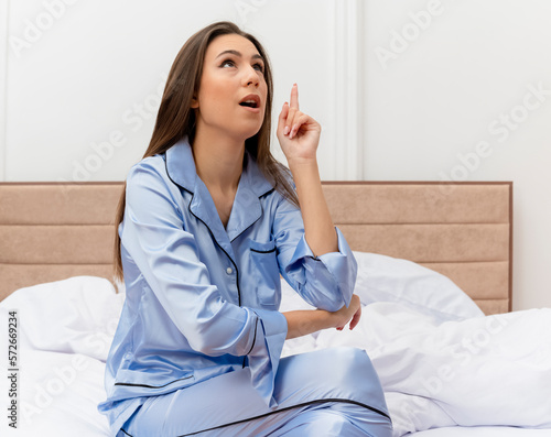 young beautiful woman in blue pajamas sitting on bed looking up amzed showing index finger having great idea in bedroom interior on light background photo