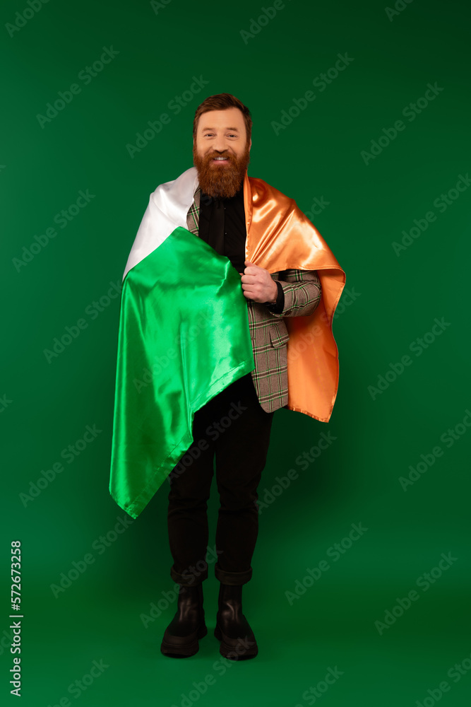 Full length of smiling bearded man wrapping in Irish flag on green background. Full length of smiling bearded man wrapping in Irish flag on green background.