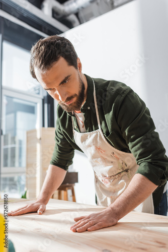 Bearded craftsman in apron touching wooden board in workshop.