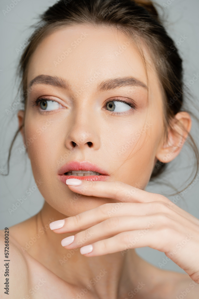 portrait of young woman with natural makeup holding hand near chin and looking away isolated on grey.