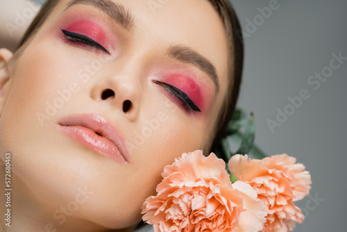 close up portrait of young woman with pink makeup posing with closed eyes near carnations isolated on grey.