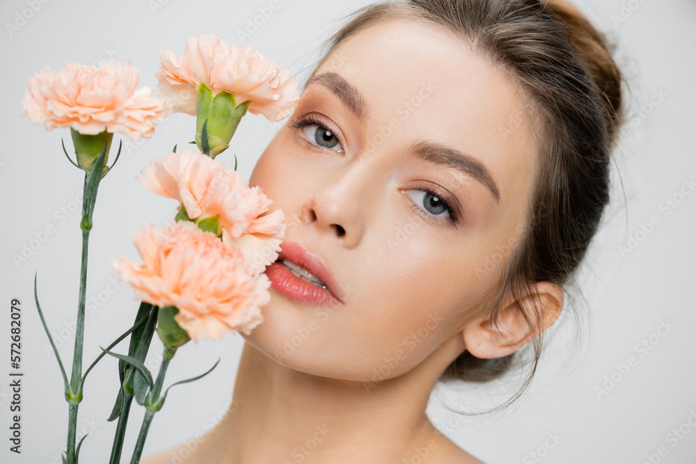 young woman with perfect skin and natural makeup looking at camera near fresh carnations isolated on grey.