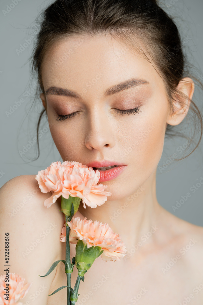 portrait of sensual woman posing with closed eyes near peach carnations isolated on grey.