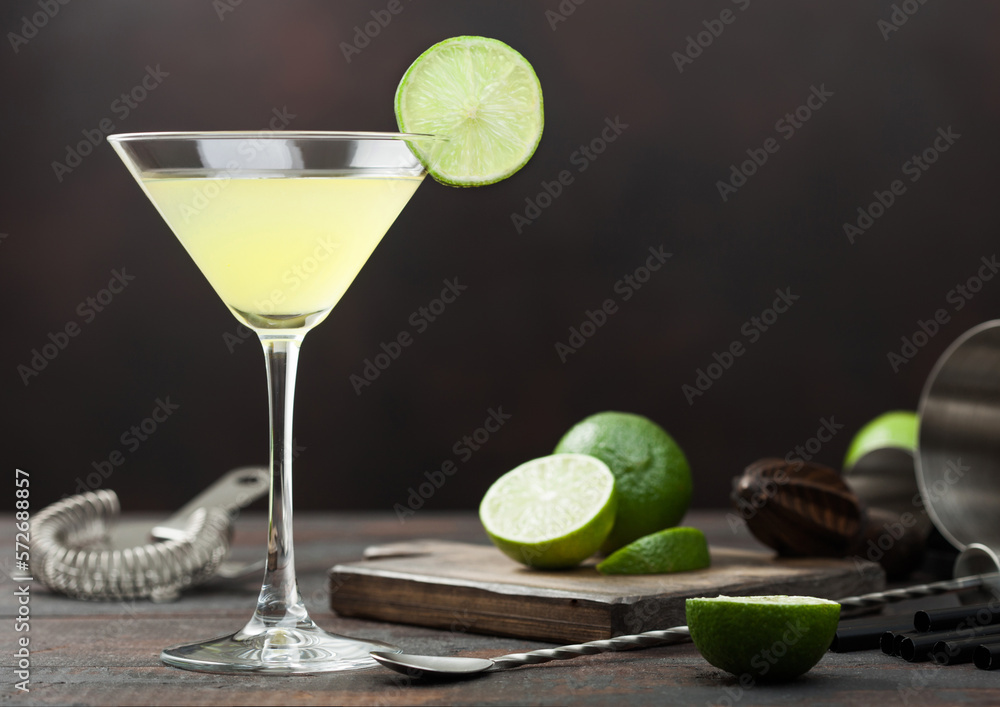 Gimlet Kamikaze cocktail in martini glass with lime slice and ice on wooden board with fresh limes and strainer with shaker.