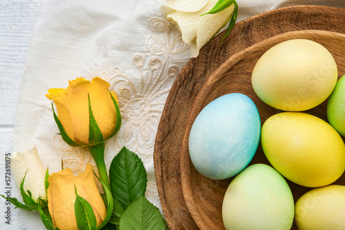 Happy Easter. Easter eggs on rustic table with white and yellow roses. Natural dyed colorful eggs in wooden plate and spring flowers in rustic room.  Toned image. Easter background with copy space.