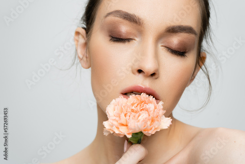 young woman with makeup and closed eyes holding peach carnation near face isolated on grey.
