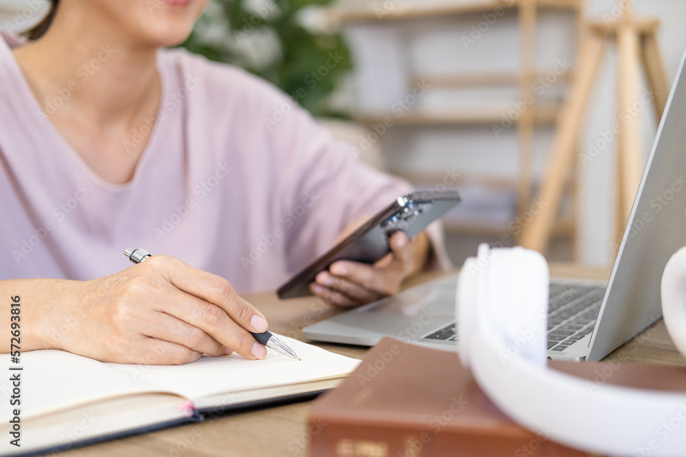 Fototapeta premium Girl college student using smartphone watching video course, zoom calling making notes in workbook sit in living room. Asian woman hold phone online learning in mobile app.