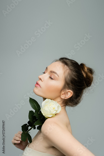sensual woman with closed eyes holding fresh rose near naked shoulder isolated on grey.