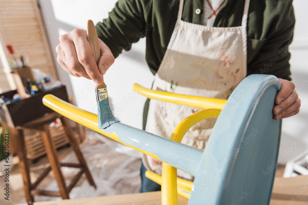 Cropped view of craftsman painting on wooden chair in blurred workshop. Cropped view of craftsman painting on wooden chair in blurred workshop.
