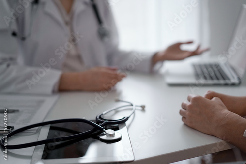 Doctor and patient sitting and discussing something at the desk in clinic. The focus is on the stethoscope lying on the table, close up. Medicine and healthcare concept