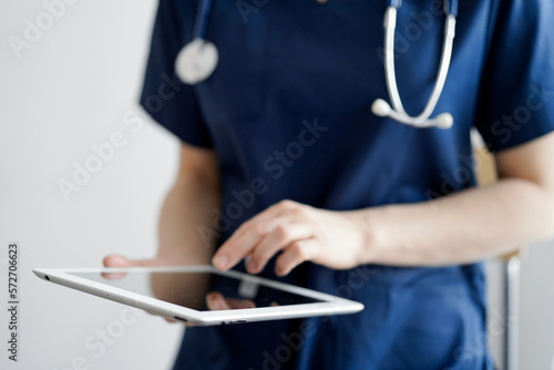 Doctor woman using tablet computer while standing near panorama window in clinic  close up. Physician or surgeon at work. Medicine concept