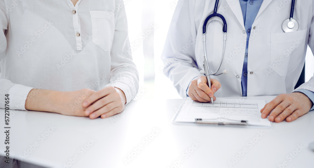 Doctor and patient sitting near of each other at the desk in clinic. The focus is on female physician's hands filling up the medication history record form or checklist, close up. Medicine concept Doctor and patient sitting near of each other at the desk in clinic. The focus is on female physician's hands filling up the medication history record form or checklist, close up. Medicine concept