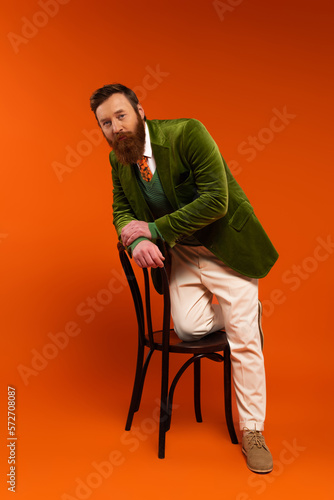 Full length of stylish bearded model posing near chair on red background.