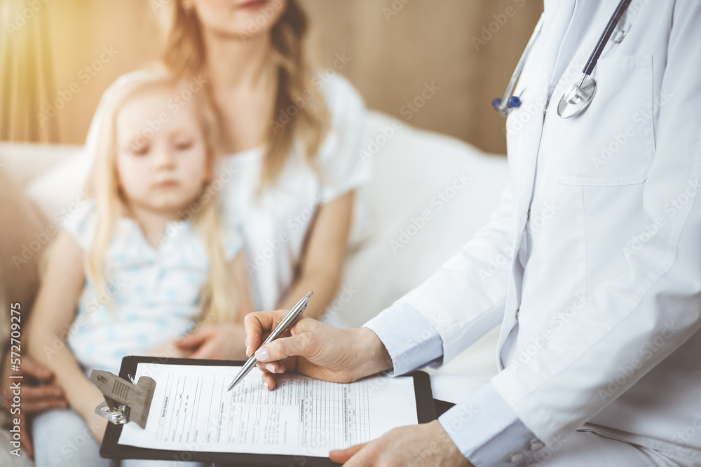 Fototapeta premium Doctor and patient. Pediatrician using clipboard while examining little girl with her mother at home. Sick and unhappy child at medical exam