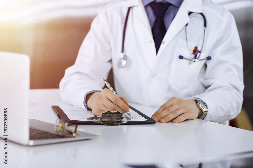 Unknown male doctor sitting and working with clipboard of medication history record in a darkened clinic, glare of light on the background, close-up of hands Unknown male doctor sitting and working with clipboard of medication history record in a darkened clinic, glare of light on the background, close-up of hands